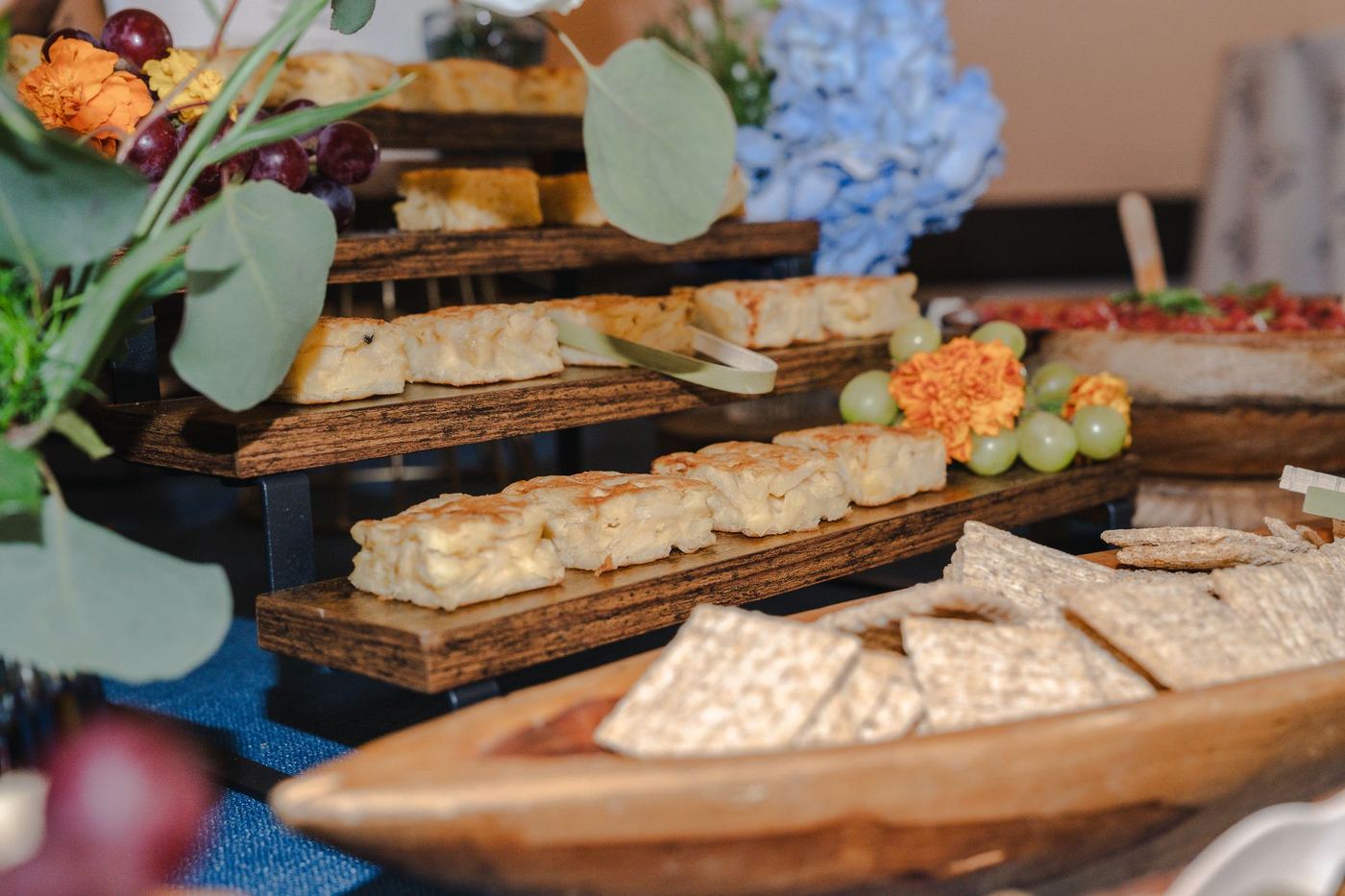 Grazing table with tiered boards of focaccia, crackers and fruit beside blue hydrangeas
