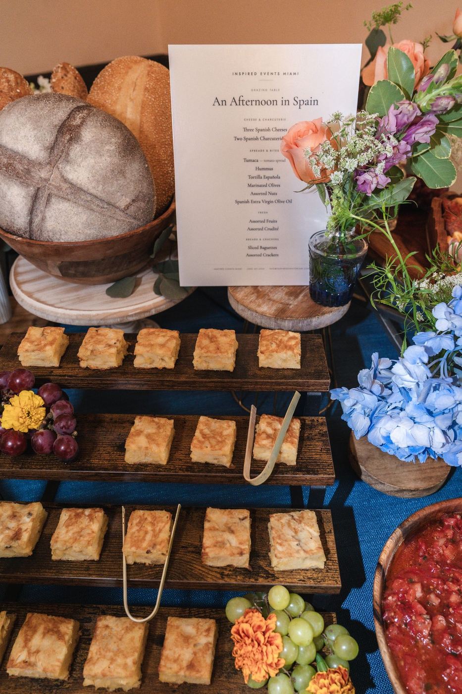 Tiered wooden risers of bread squares with a menu card, flowers and a bowl of tomato