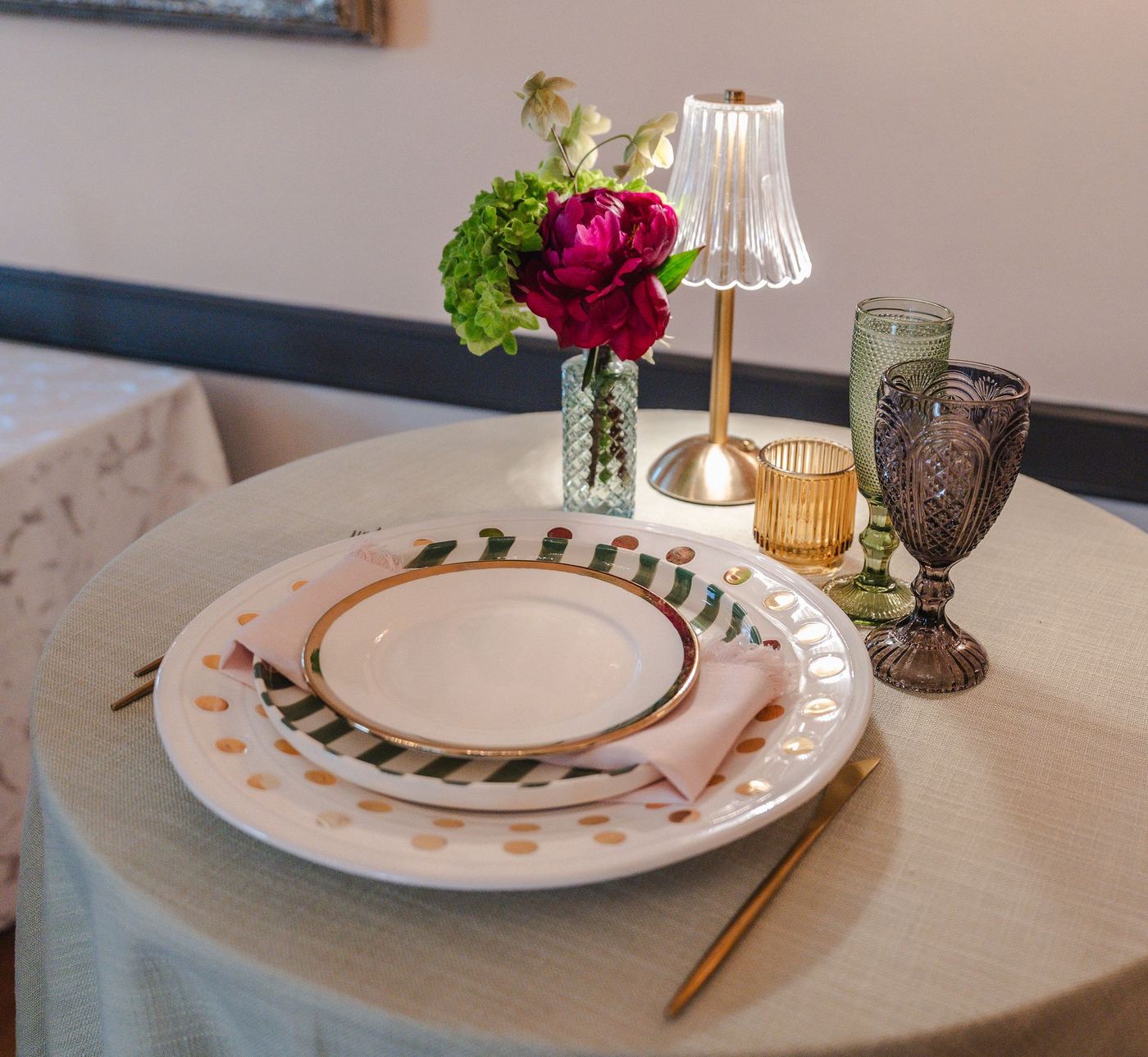Round table in matcha green Expo with an Aubrey Golden Glam charger, a striped Juno plate, a Halo Gold plate and a blush Alegria Fringe napkin